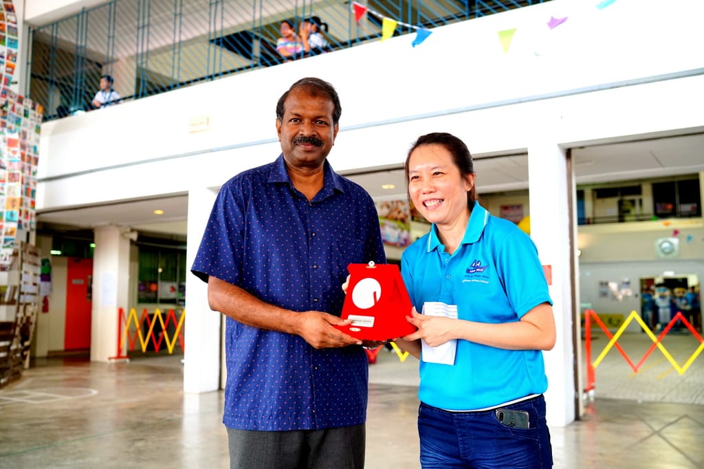 A man in a blue shirt and a woman in a teal polo together hold a red gift inside a building decorated with colorful flags.